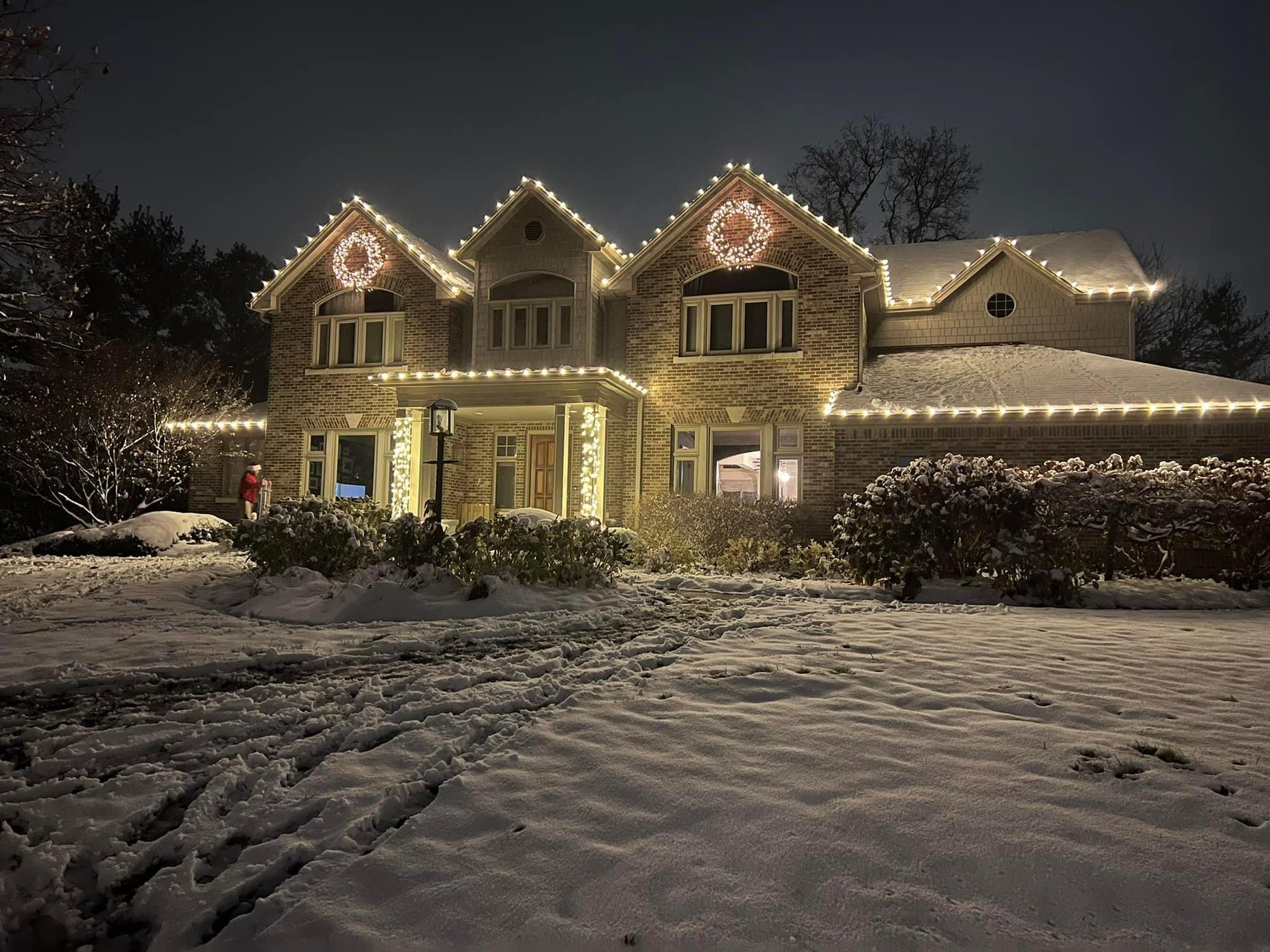 Stone and brick Indiana home lit with warm white Christmas lights along the rooflines and gable wreaths, with snow across the front lawn