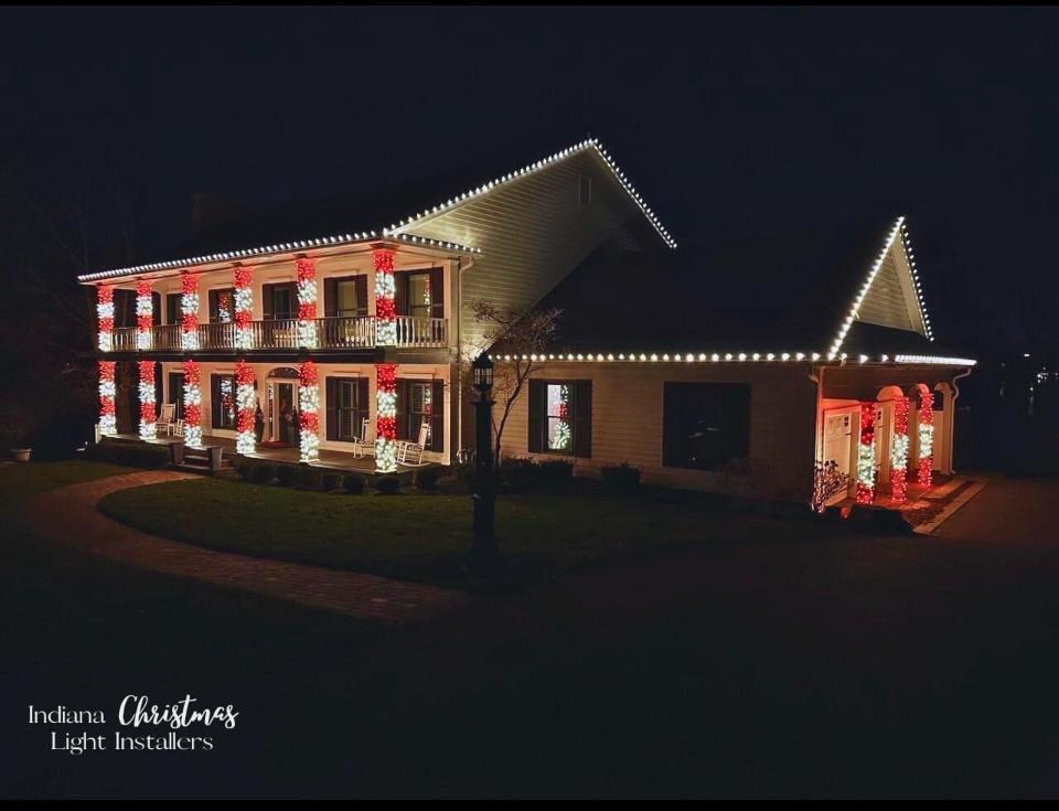 Two story white Indiana home with warm white Christmas lights along every roofline and all six porch and balcony columns wrapped in red and white lights, with a lit entry and lamp post