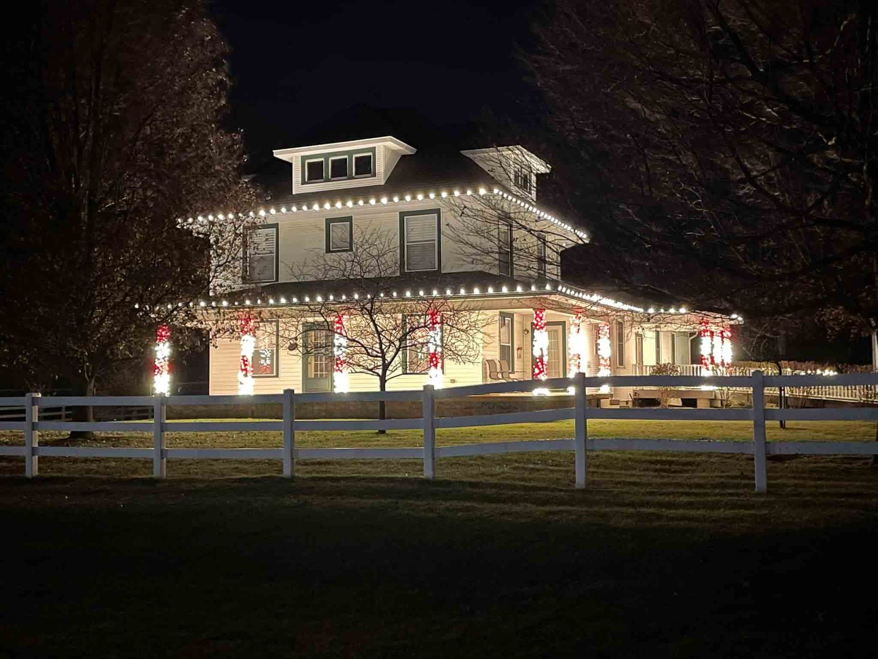 White Indiana farmhouse behind a rail fence, outlined in warm white Christmas lights with red bows