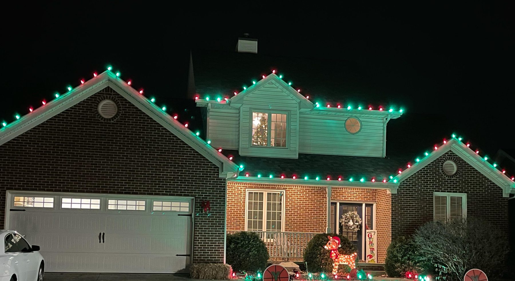 Indiana home outlined in red and green Christmas lights with lit shrubs, a lighted reindeer and red and green lights along the front garden
